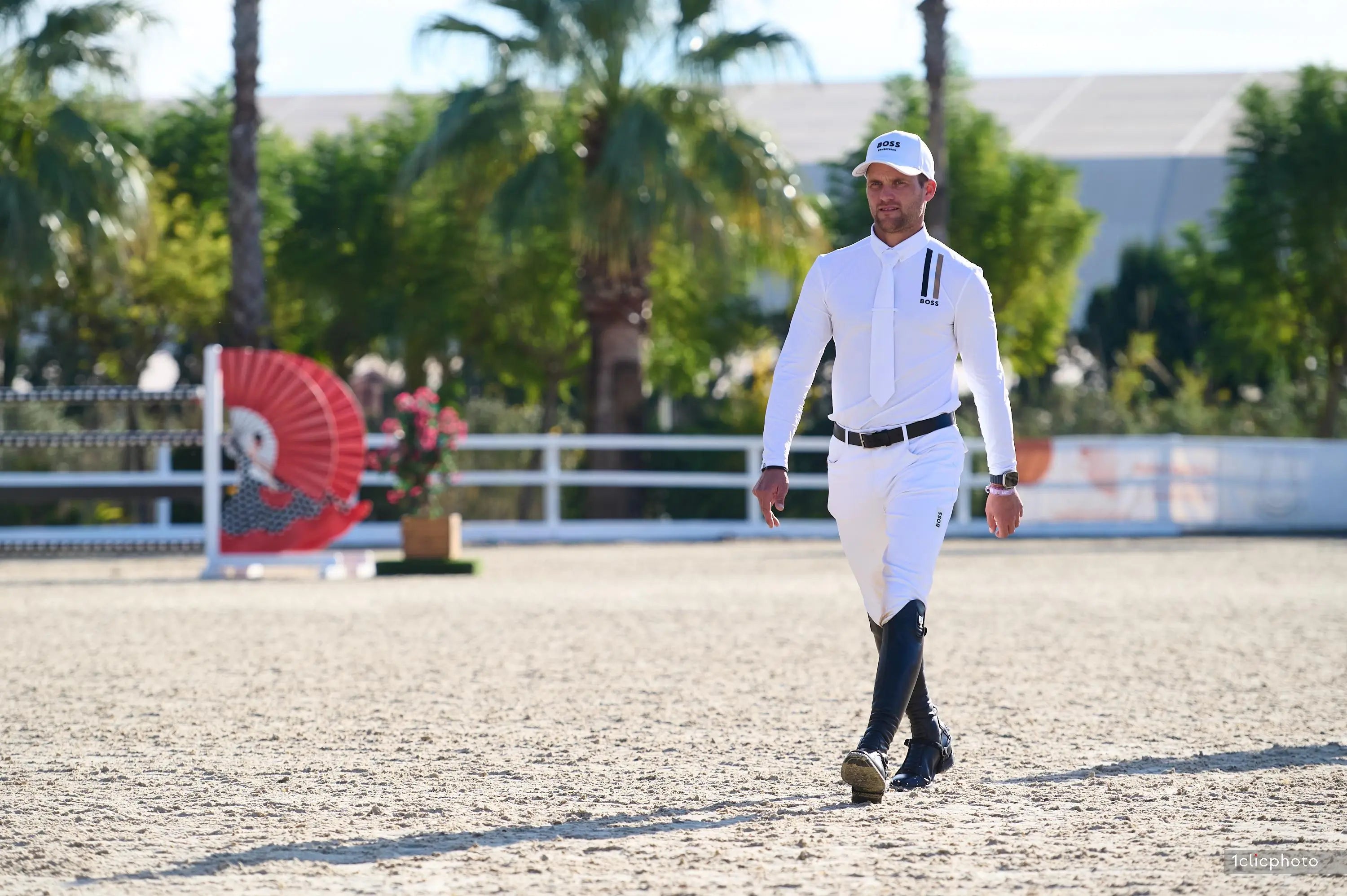 Alexander Schill in equestrian attire walking on a dirt path with palm trees in the background