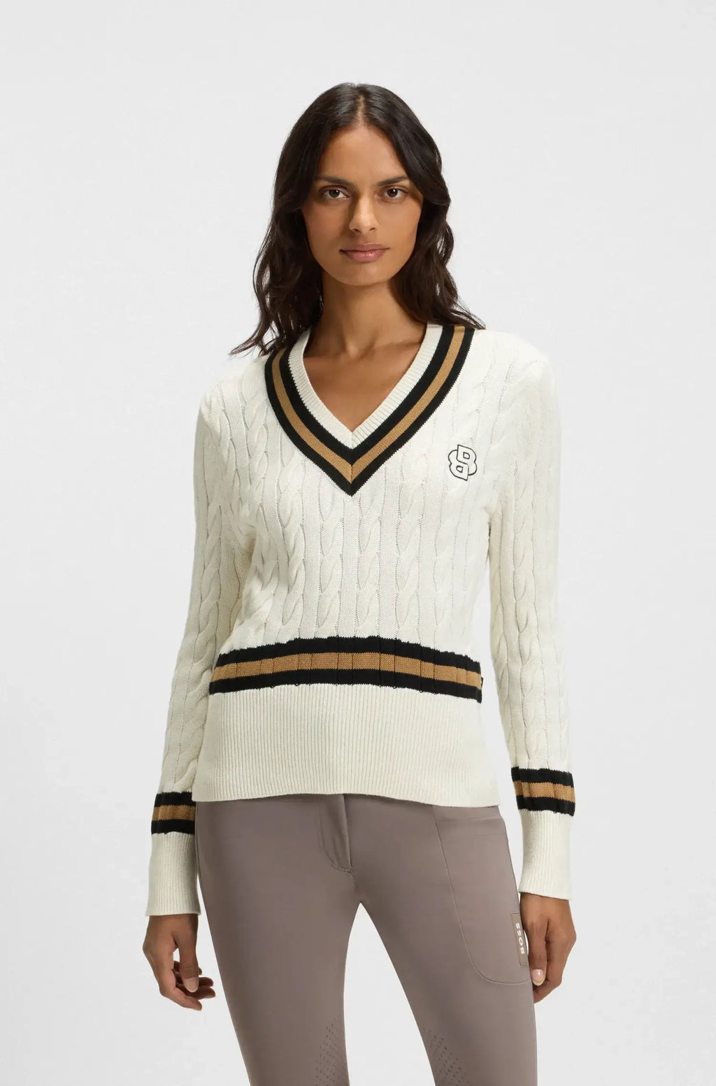 Soft Creme | A woman modeling a cream cable-knit V-neck sweater with black and tan stripes on the collar and hem, paired with taupe pants, against a light gray studio background.